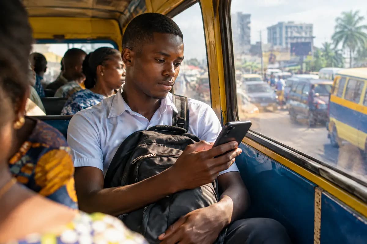 Un étudiant, sac sur les genoux, consulte son téléphone près de la fenêtre d’un minibus dans la circulation de Kinshasa.