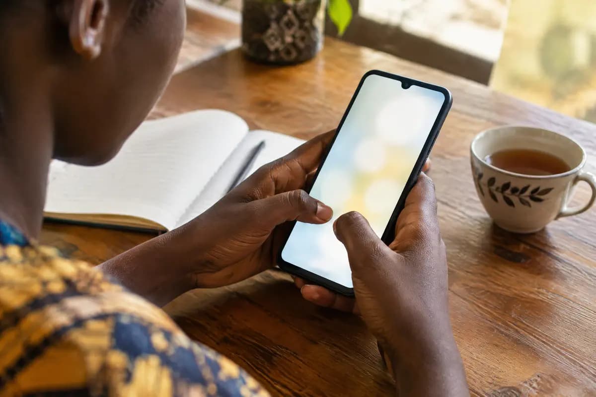 Gros plan sur des mains qui tapent une note sur un téléphone, à côté d’un carnet ouvert et d’une tasse de thé.